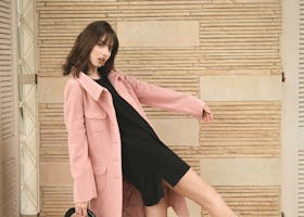 Fashion model in pink coat and black dress striking a dynamic pose indoors with stylish black heels.